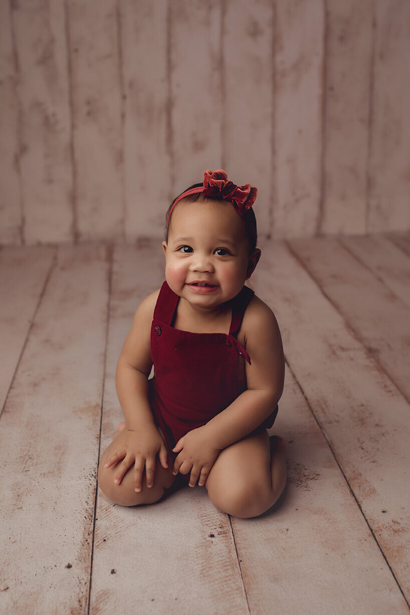a one year old girl sitting on her knees in a burgandy romper on a wood backdrop