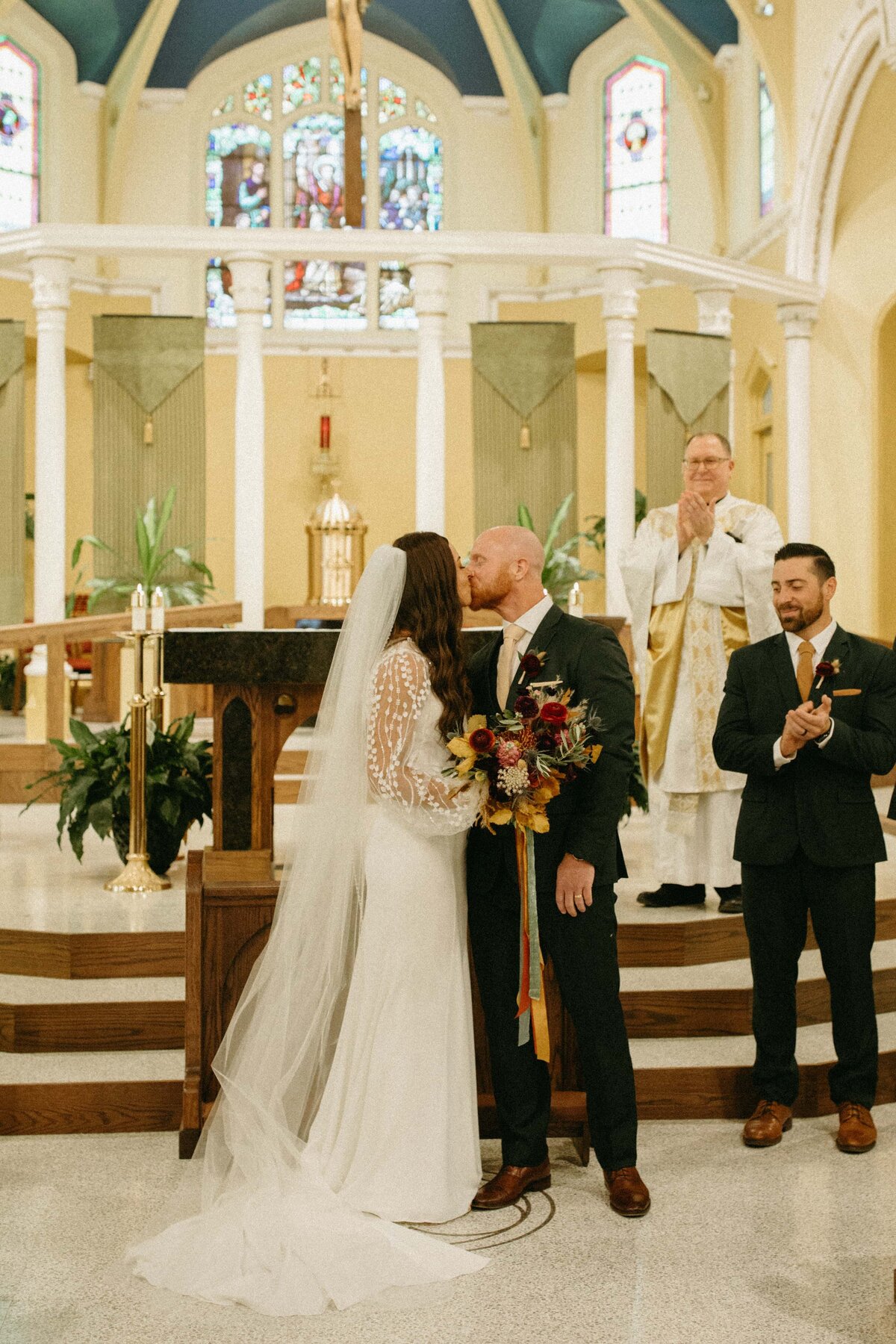 A bride and groom kiss at the altar of a church during their wedding ceremony, with a priest and a wedding coordinator watching in the background.