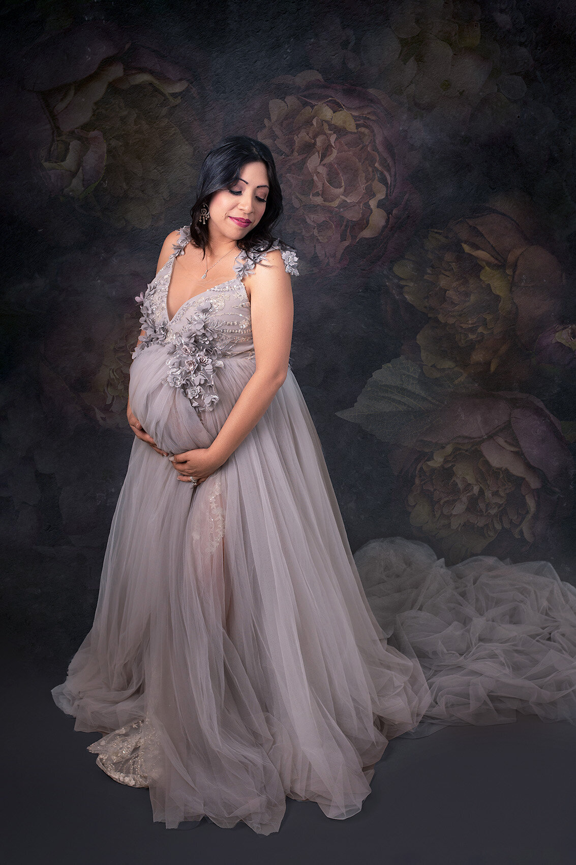 Pregnant woman in a couture gown by Before And Ever, standing in front of a floral backdrop.