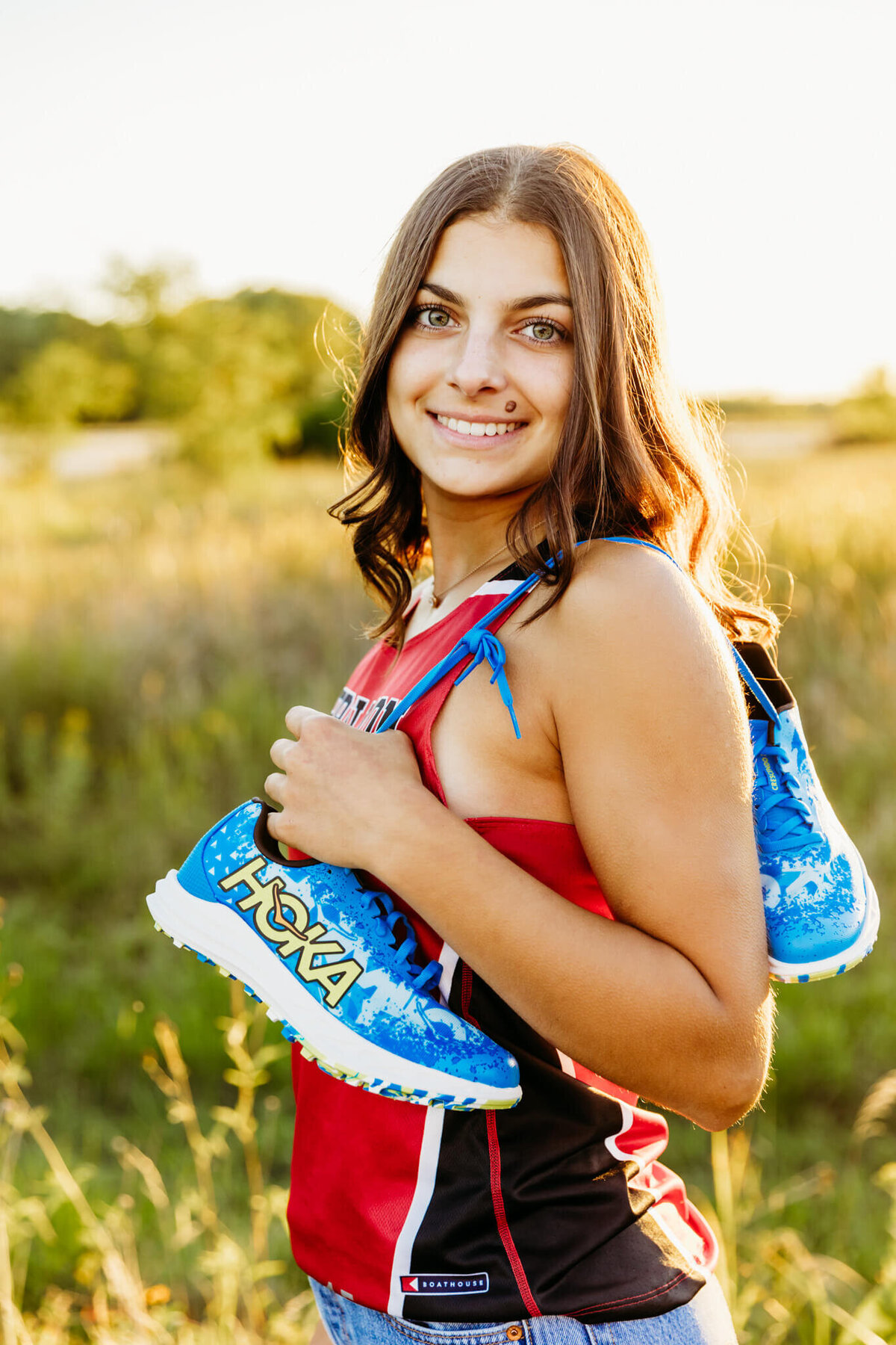brunette teen girl in  her red and black track jersey holding her blue track shoes over her shoulder and smiling