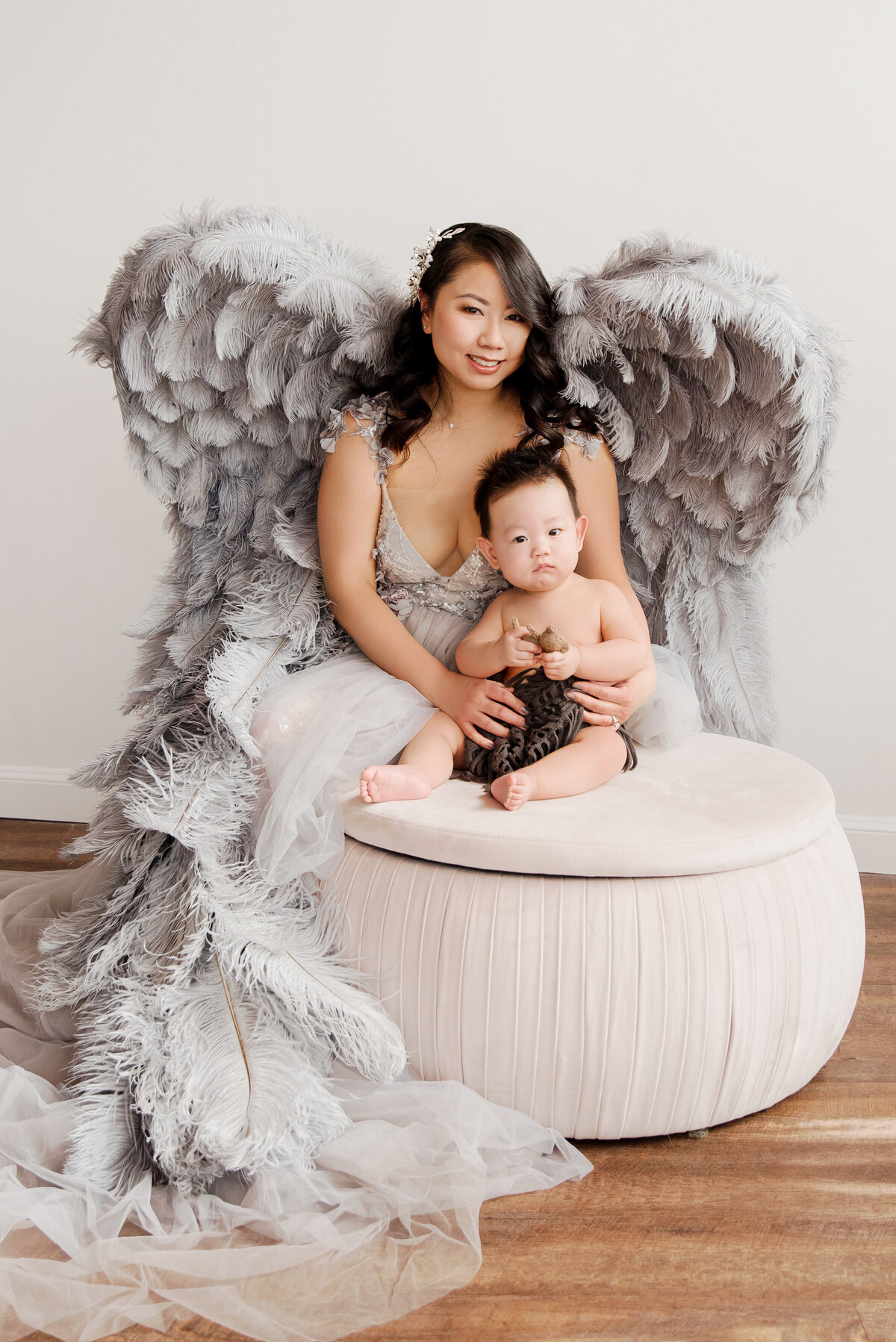 st-louis-motherhood-photographer-mother-in-silver-gown-with-silved-angel-wings-sitting-behind-baby-on-poof