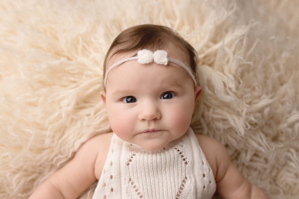 Six Month baby girl dressed in white romper with matching bow laying on white flokati rug