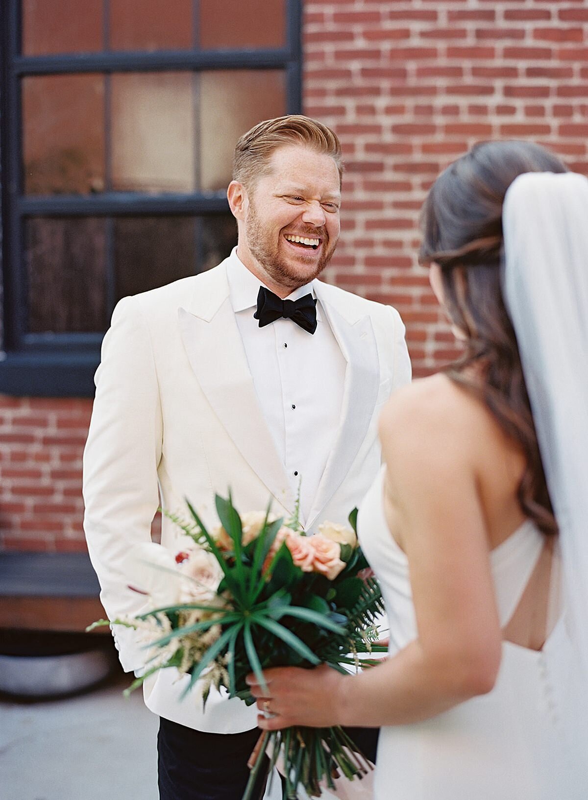 The groom, wearing a tuxedo with a white jacket laughs at something the bride, wearing a white silk halter top sheath wedding dress and veil, says to him. The bride holds her tropical bouquet of palm fronds, pink, peach, yellow, white and ivory flowers and she laughs with the groom.