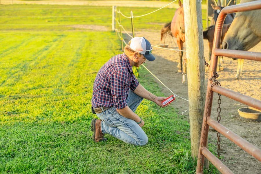 A farmer in a plaid shirt and cap inspecting or repairing a fence with a tool in a pasture, with a horse partially visible in the background.