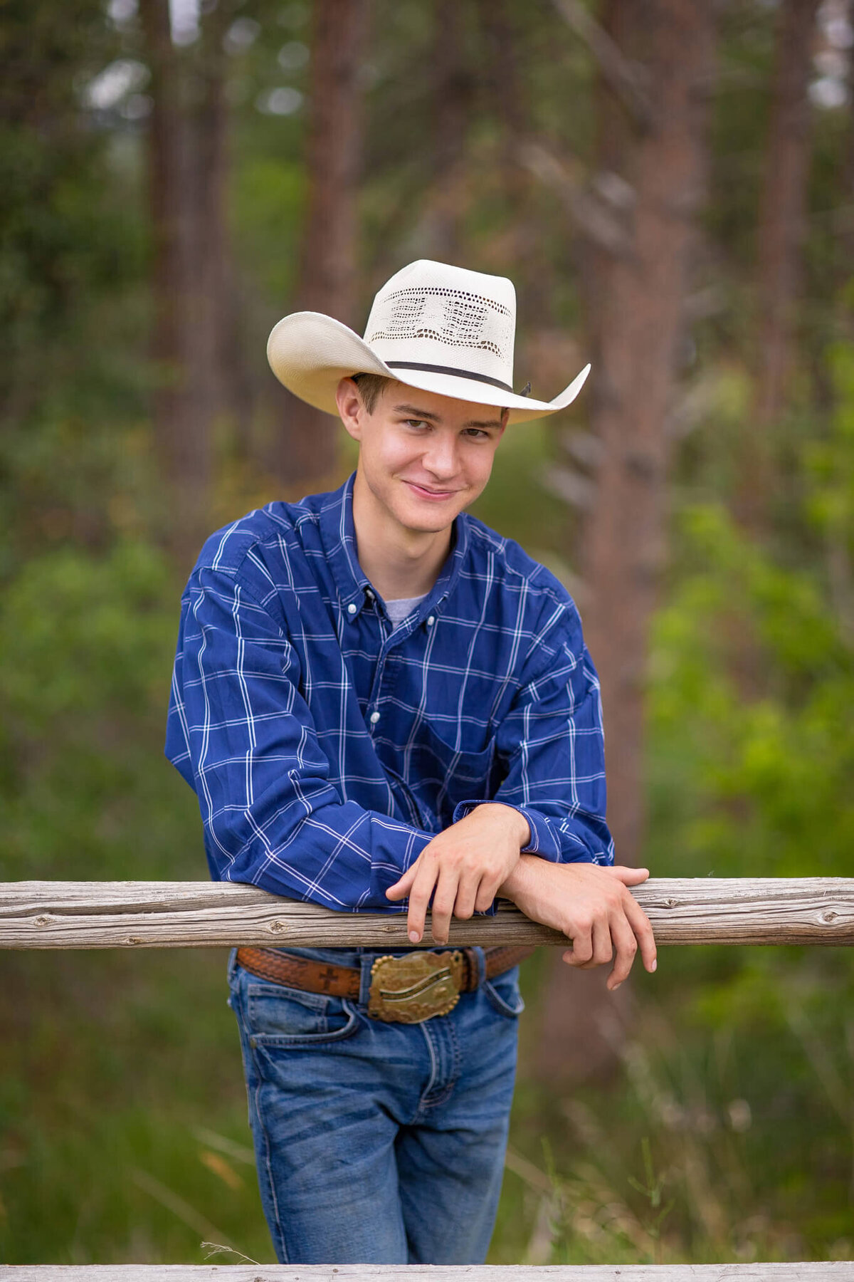 teenage boy in a blue shirt and jeans wearing a cowboy hat leaning against a fence