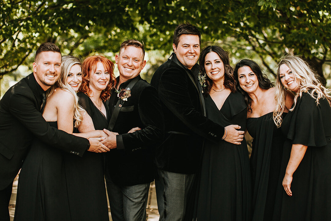 Two grooms wearing black tuxedos share smiles outside with their wedding party also wearing all black.
