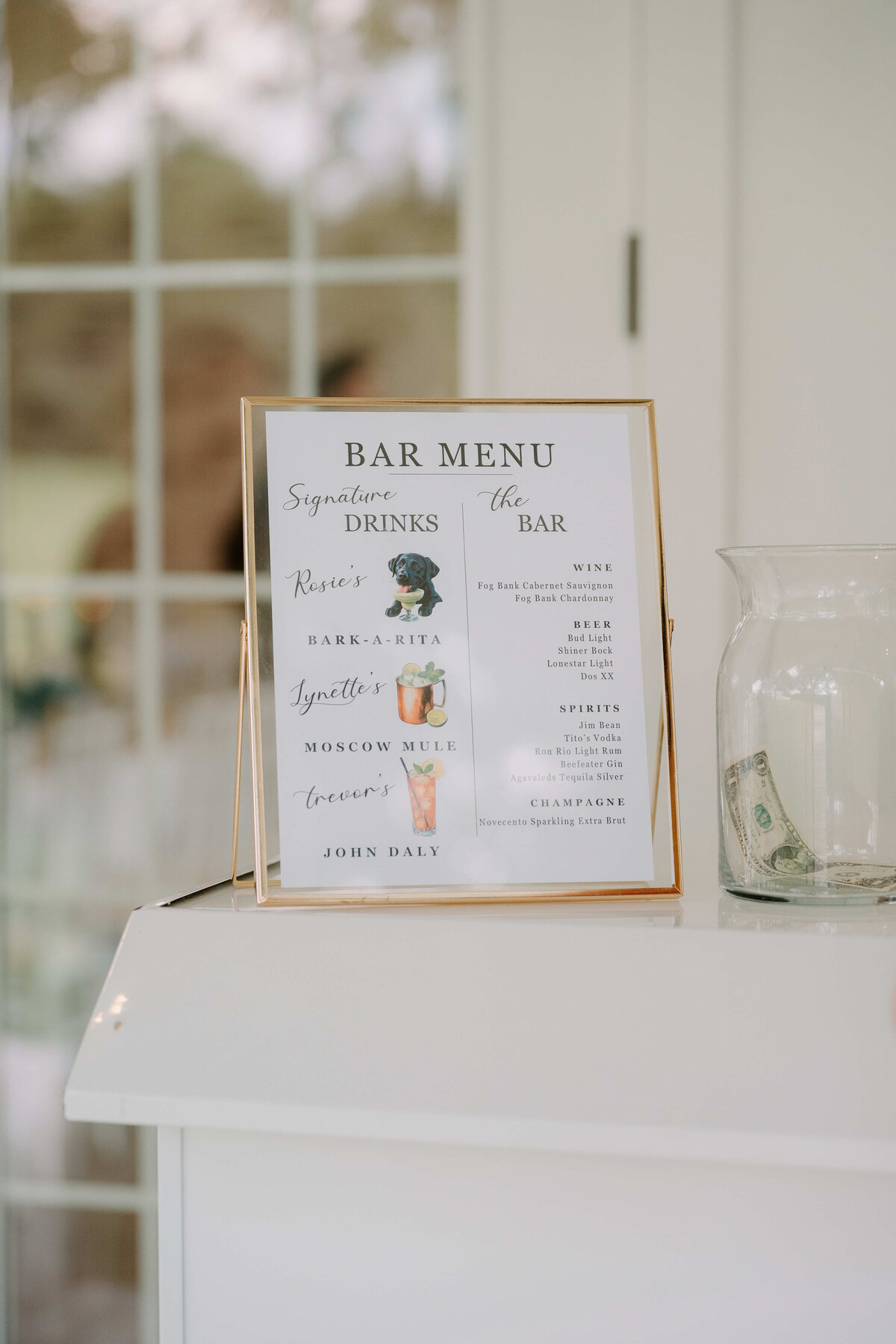 Tabletop bar menu in a gold frame listing signature drinks, with images of cocktails. A tip jar with cash is beside it. The setting is bright and elegant.