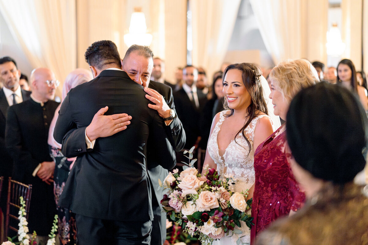A wedding scene with a smiling bride holding a bouquet of flowers standing next to her family. Two men are embracing warmly. The guests in formal attire are observing the event in an elegantly decorated venue.