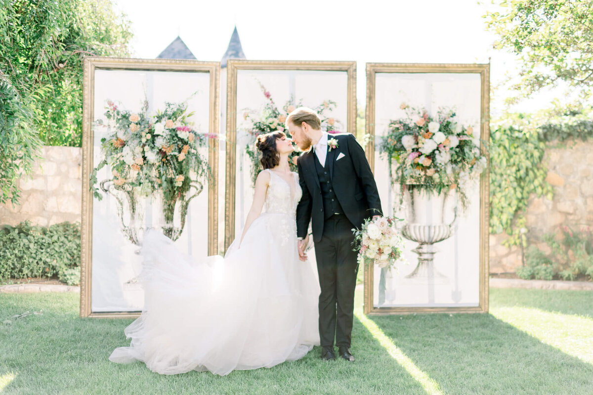 bride in white wedding gown and groom in black suit kiss in front of three panels of faux planters with real flowers