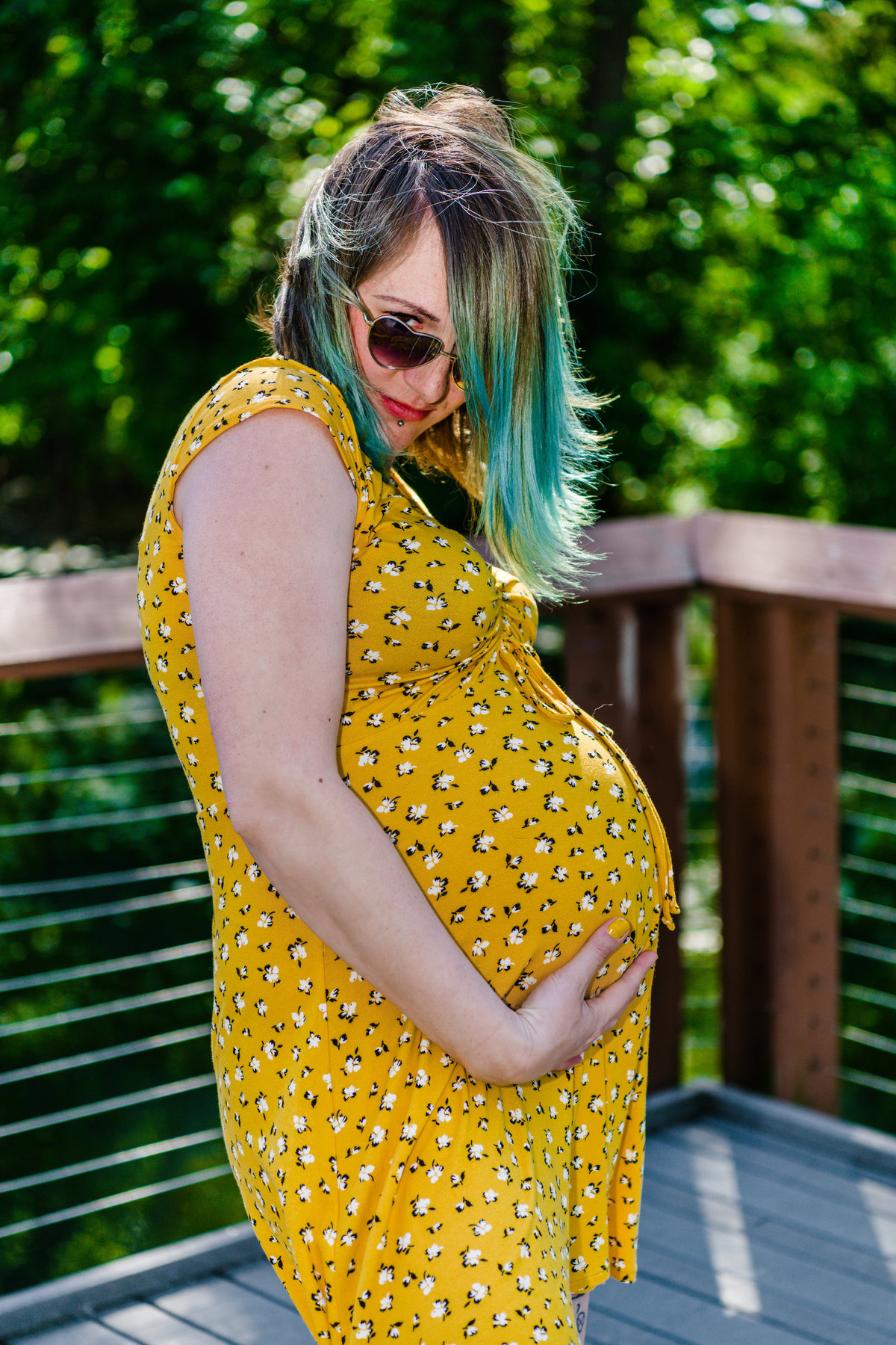 Pregnant woman with green hair and sunglasses holds stomach.