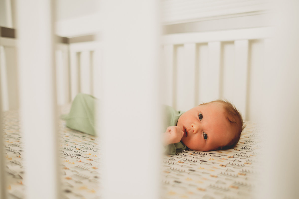 newborn baby boy peeking through white crib slats in nursery