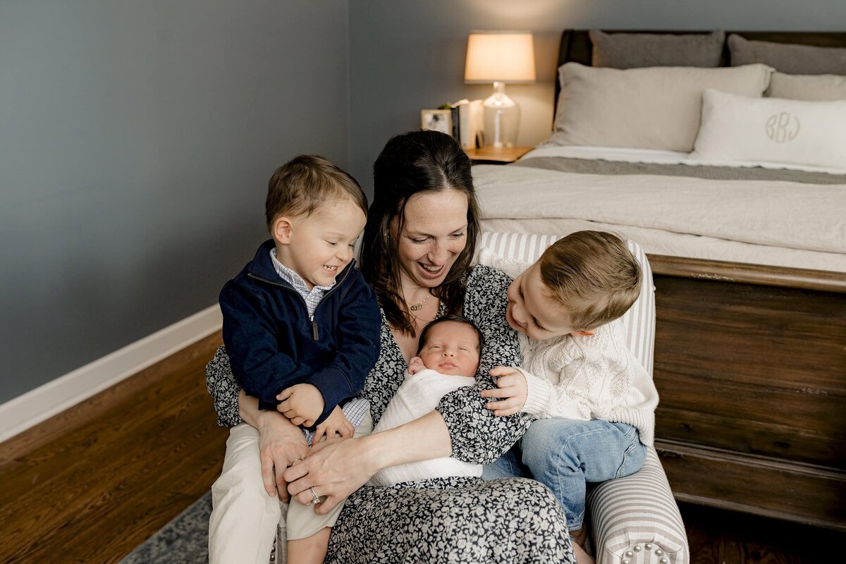 mother sits in her bedroom with all three of her sons around her during their lifestyle newborn session in their charlotte home