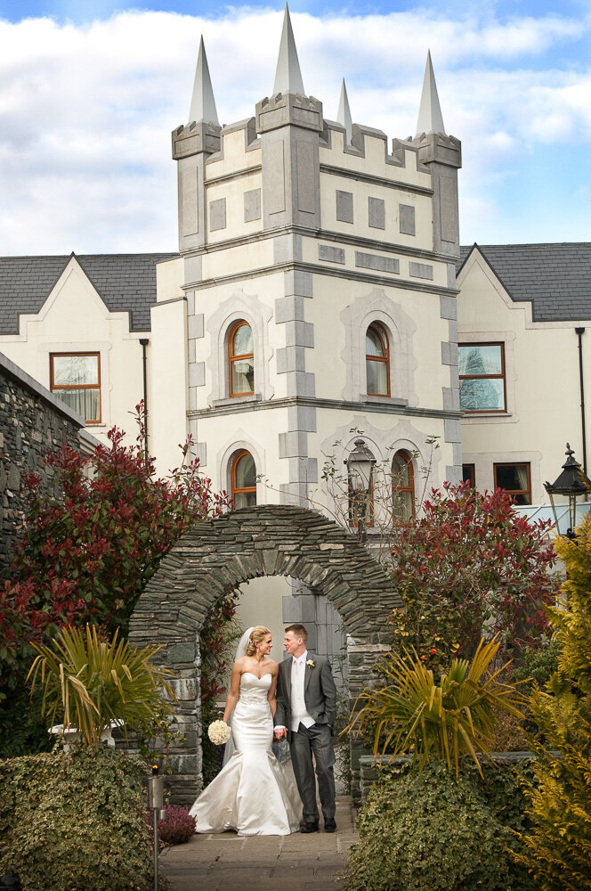 Bride in satin, mermaid style wedding dress, holding a white bouquet while walking with her groom in a grey tailcoat and white tie at the Muckross Park Hotel in Kerry