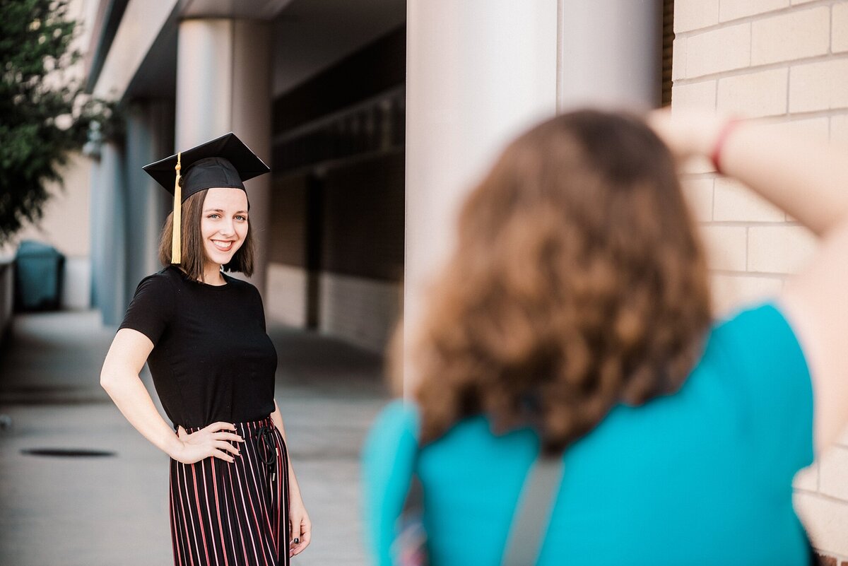 A college graduate puts a hand on her hip amongst some columns while a photographer takes her picture