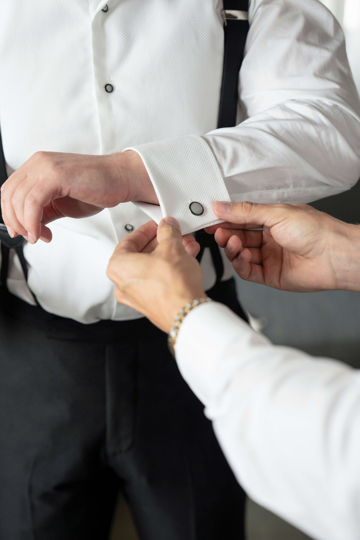 A person assists another with fastening cufflinks. Both are wearing formal attire with white shirts.