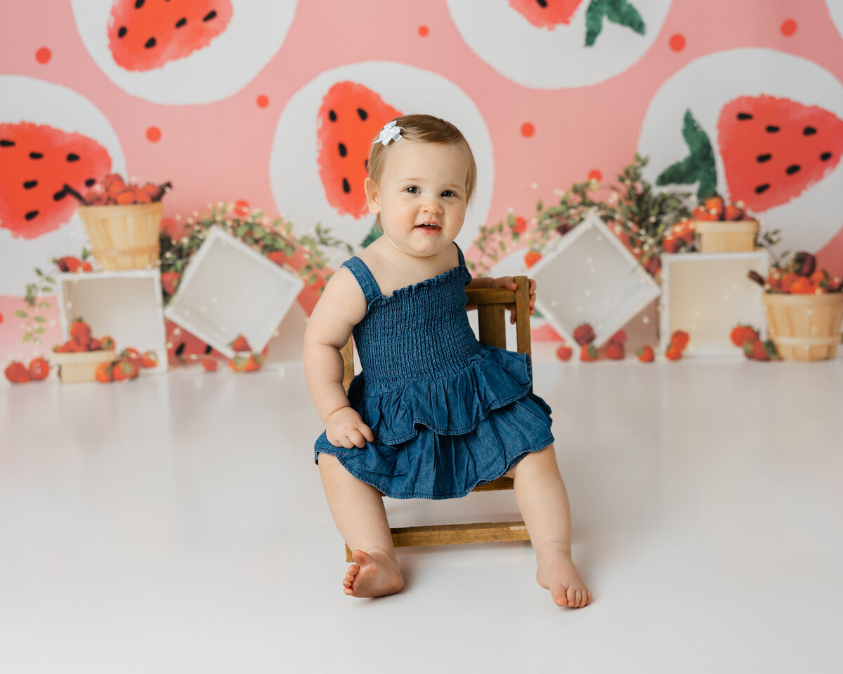 Charming one-year-old girl sitting on a small chair, wearing a sweet denim dress, with a playful smile against a vibrant strawberry-themed backdrop. This joyful moment was captured by Yucaipa cake smash photographer Kaitlyn Dawn Photography