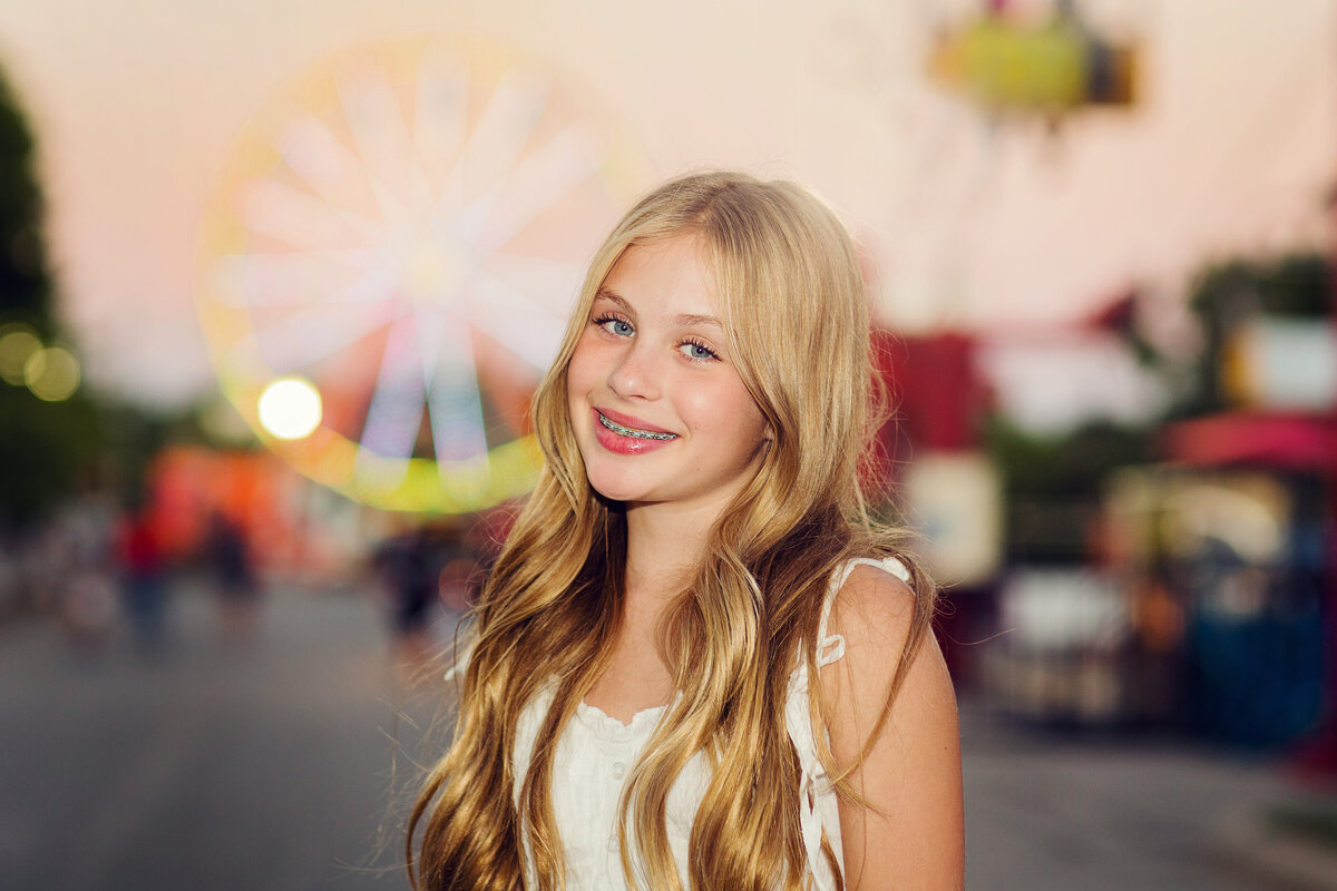 Tween girl with long, blonde hair is smiling at the camera with the ferris wheel behind her at the Indiana State Fair.