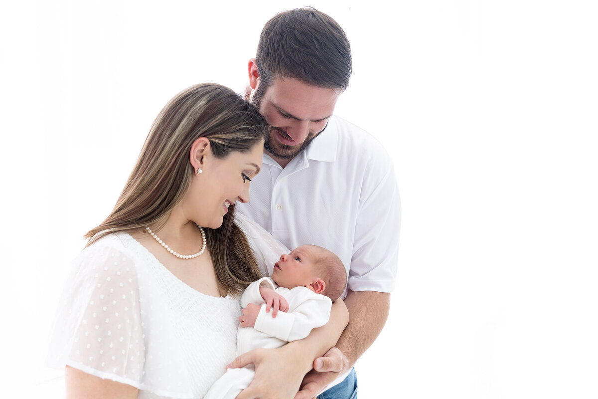 Happy new parents smile down at their awake newborn baby in mom's arms while standing in the studio of an Atlanta newborn photographer