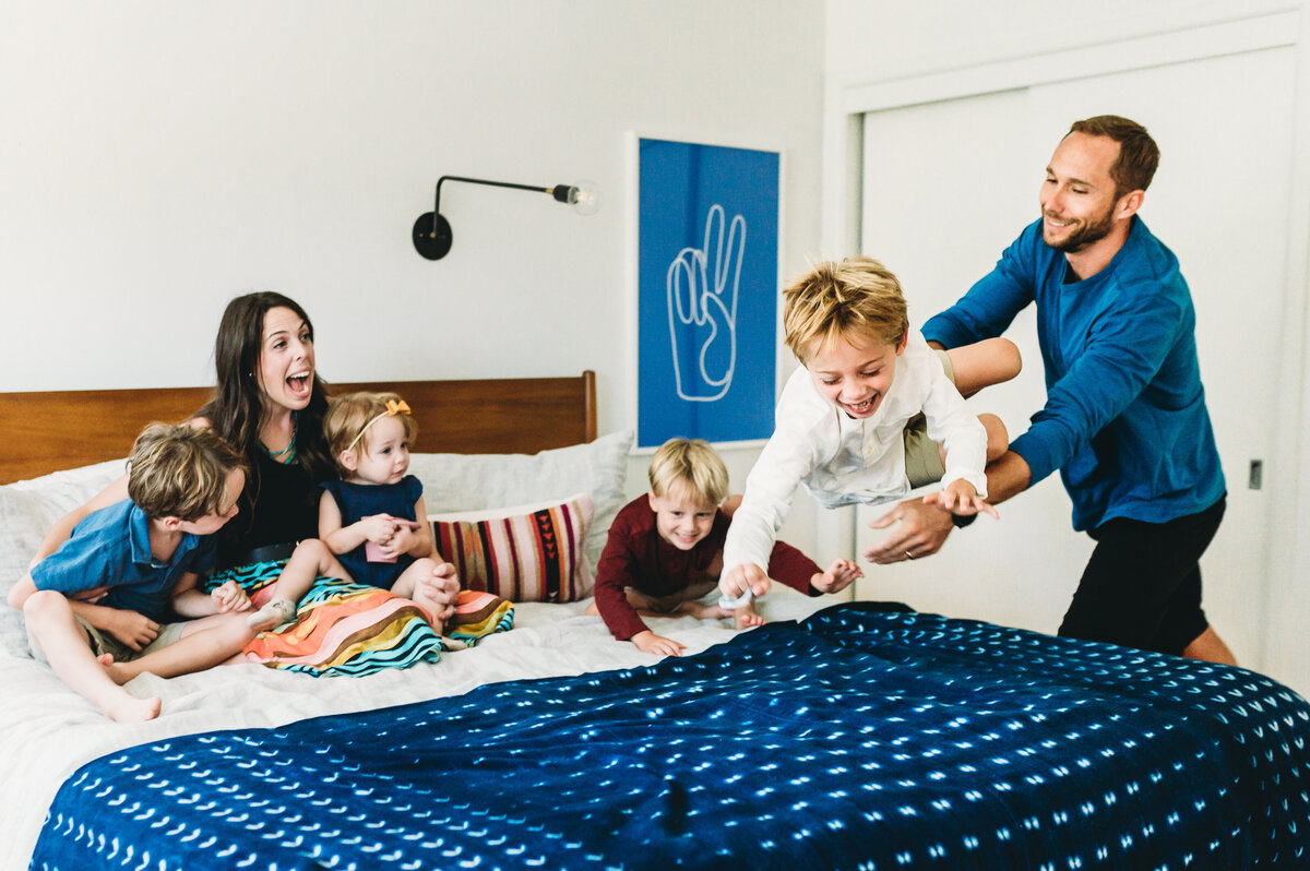 family of 6 in a bedroom laughing with dad tossing son on bed