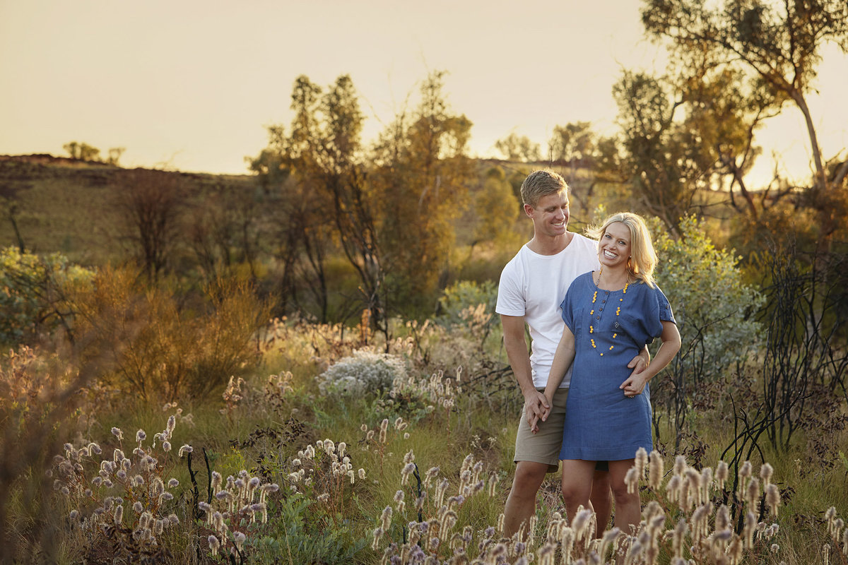 Husband looking at wife and wife looking at camera smiling with golden sunlight streaming through and natural bush and mountains in the background