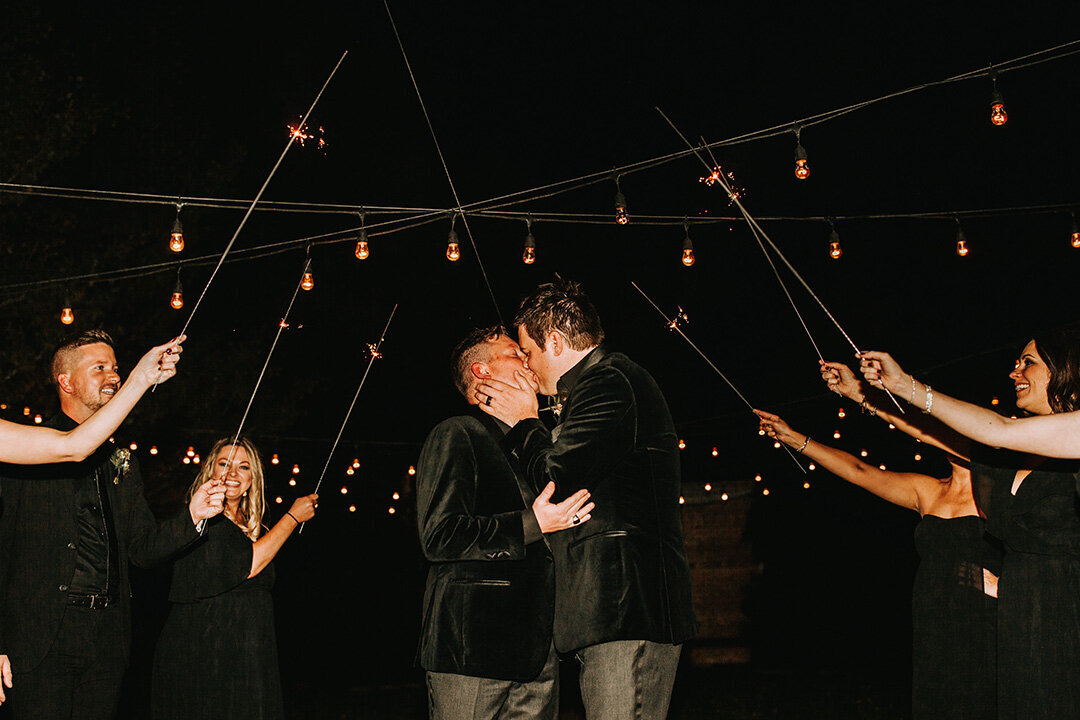 Two grooms wearing black tuxedos kiss at night, while guests wearing all black hold sparklers above.