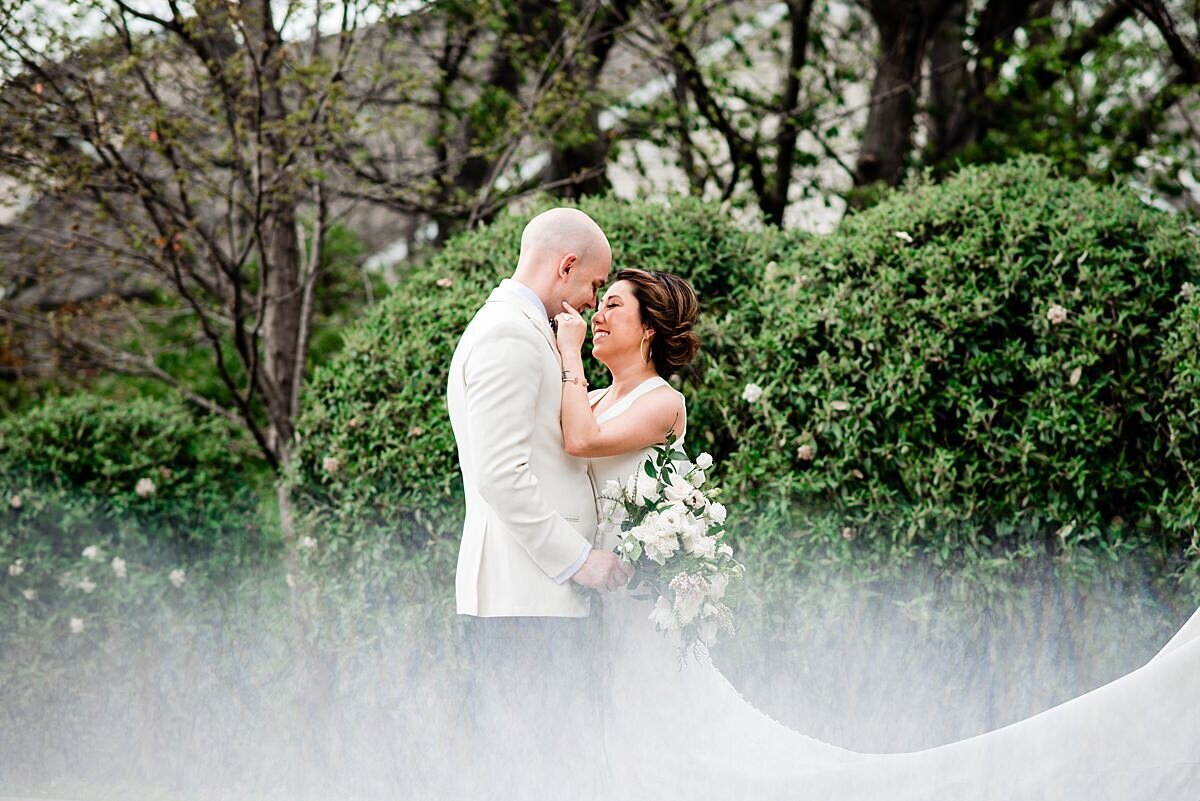 The bride and groom stand in the misty forest and they embrace at Noah Liff Opera Center. The groom is wearing a tuxedo with a white jacket and black pants. The bride is wearing a fitted white dress with a long train. She is holding his cheek while he holds her white floral bouquet.