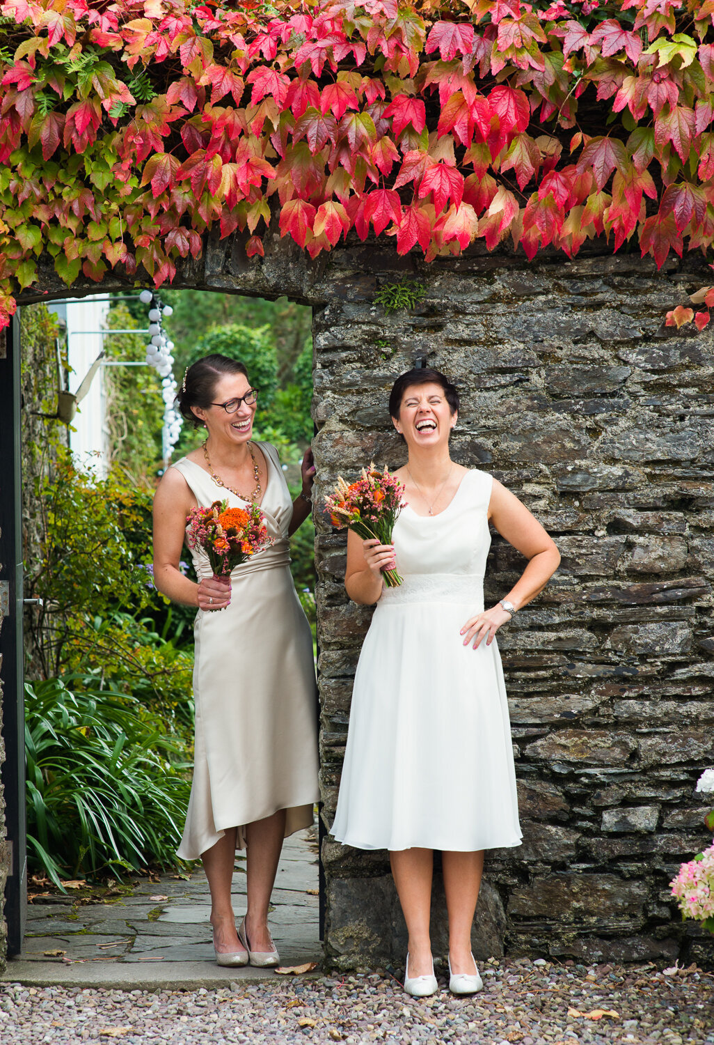 Gay brides holding orange flower bouquets, laughing underneath red and orange ivy leaf