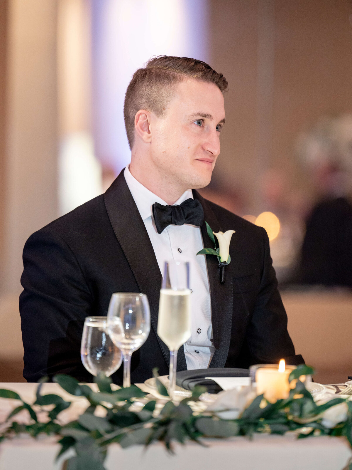 A person in a black tuxedo with a white shirt and bow tie sits at a table set with a candle, a wine glass, and a champagne flute. The table is decorated with greenery, and the person has a small boutonniere on their jacket.