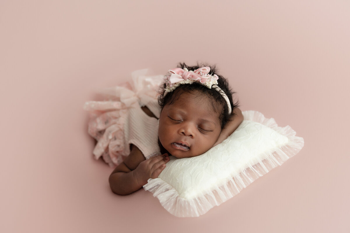 A newborn baby girl in a pink dress and headband sleeps on her belly on a white pillow
