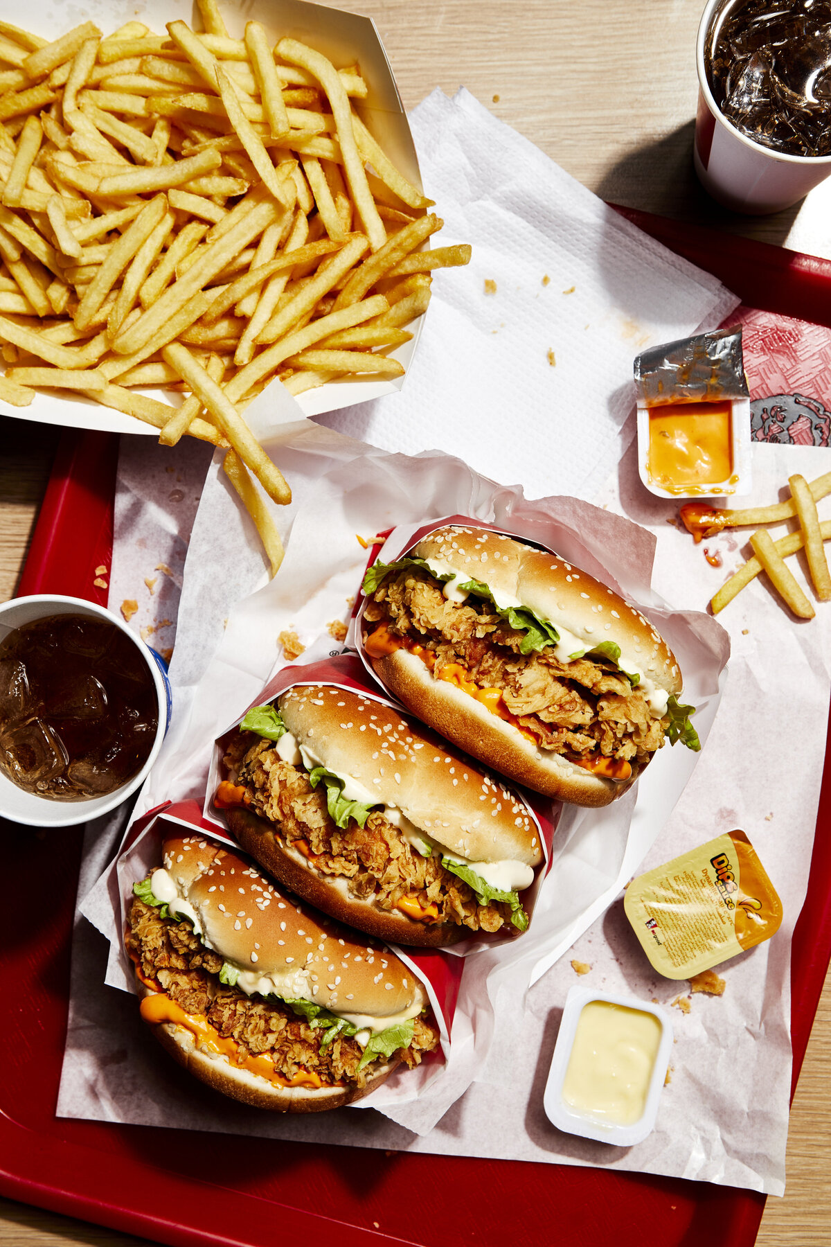Three chicken sandwiches on a tray with fries, drinks, and dipping sauces.