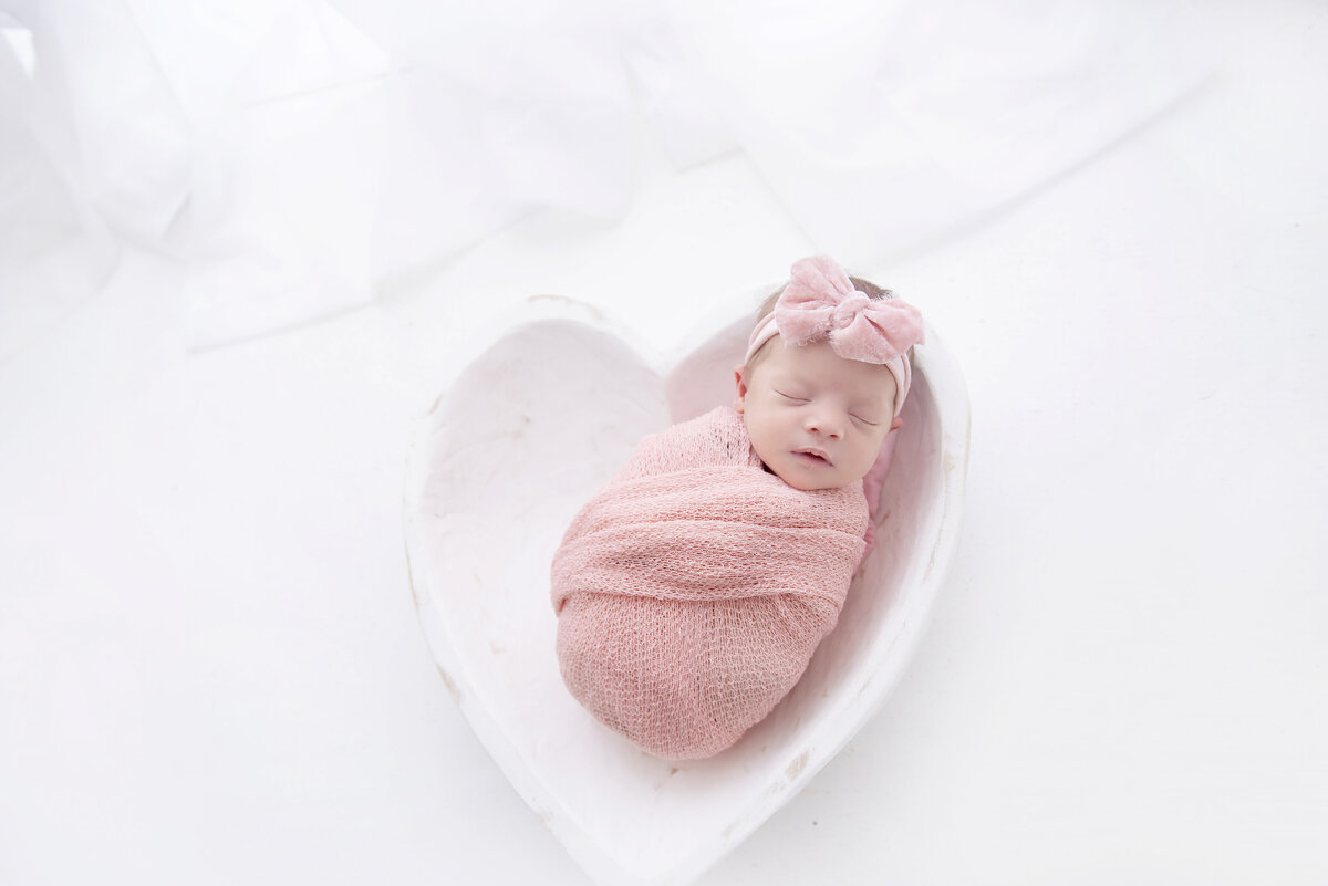 A newborn baby girl sleeps in a pink swaddle and matching bow in a heart shaped bowl