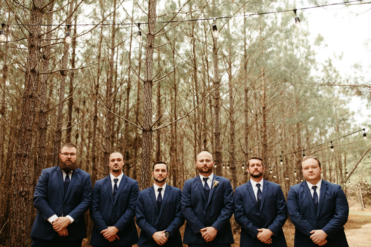 Groom and groomsmen in blue suits posing in the woods with string lights hanging above them