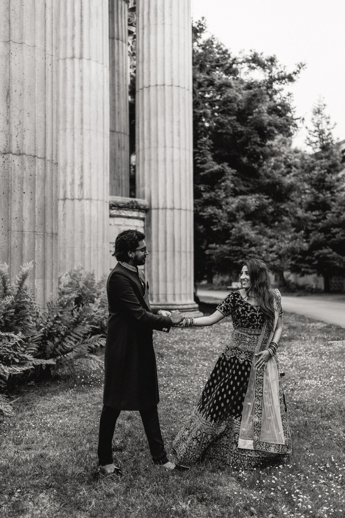 A black and white photograph of Heera and Nithin during their engagement portrait session at the Palace of Fine Arts in San Francisco, California. They are dressed in traditional clothing from India and are holding hands as they walk together. Heera is leading slightly and smiling as she looks back at Nithin. Wedding photographs by Stacie McChesney/Vitae Weddings.