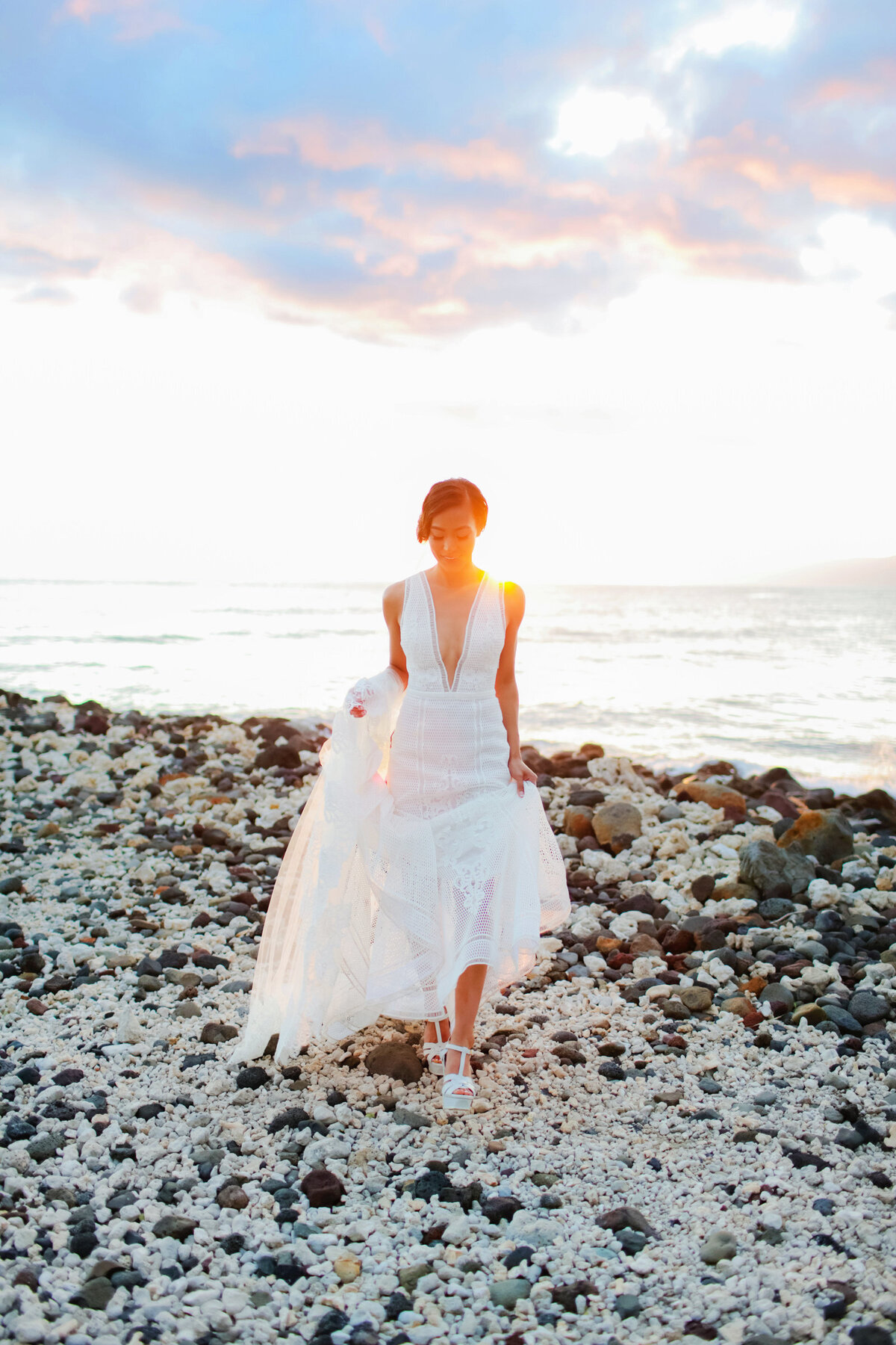 Beautiful Asian bride holds the hem of her wedding gown and walks in heels over lava rock on the island of Maui