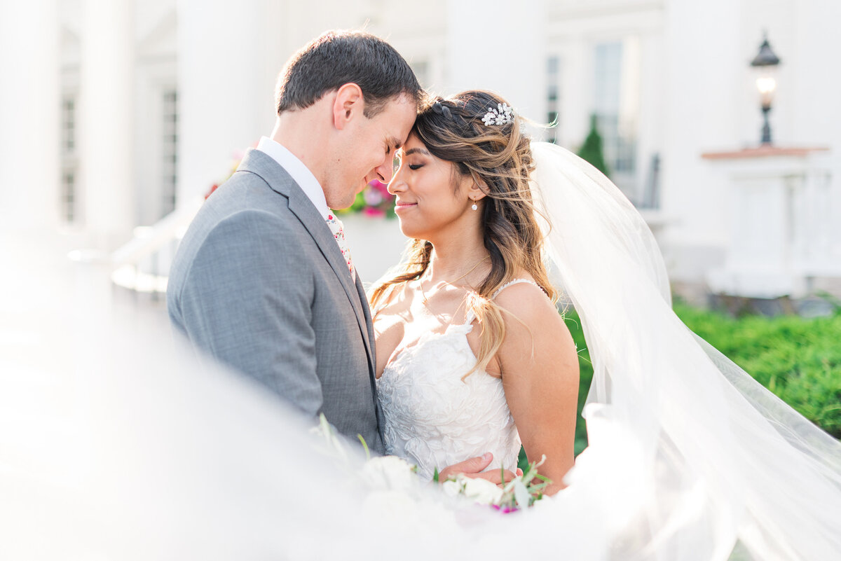 Groom and bride standing forehead to forehead with bride's veil wrapping around them in front of a white manor house.