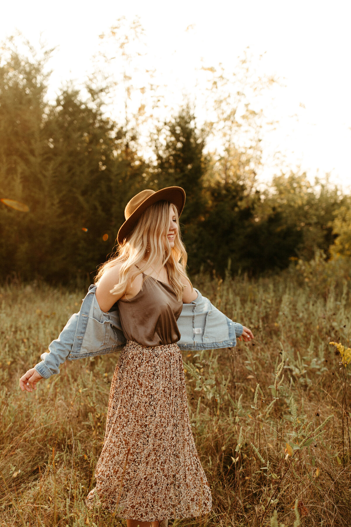 High school senior in dress with boho hat and denim jacket twirling in a field at golden hour