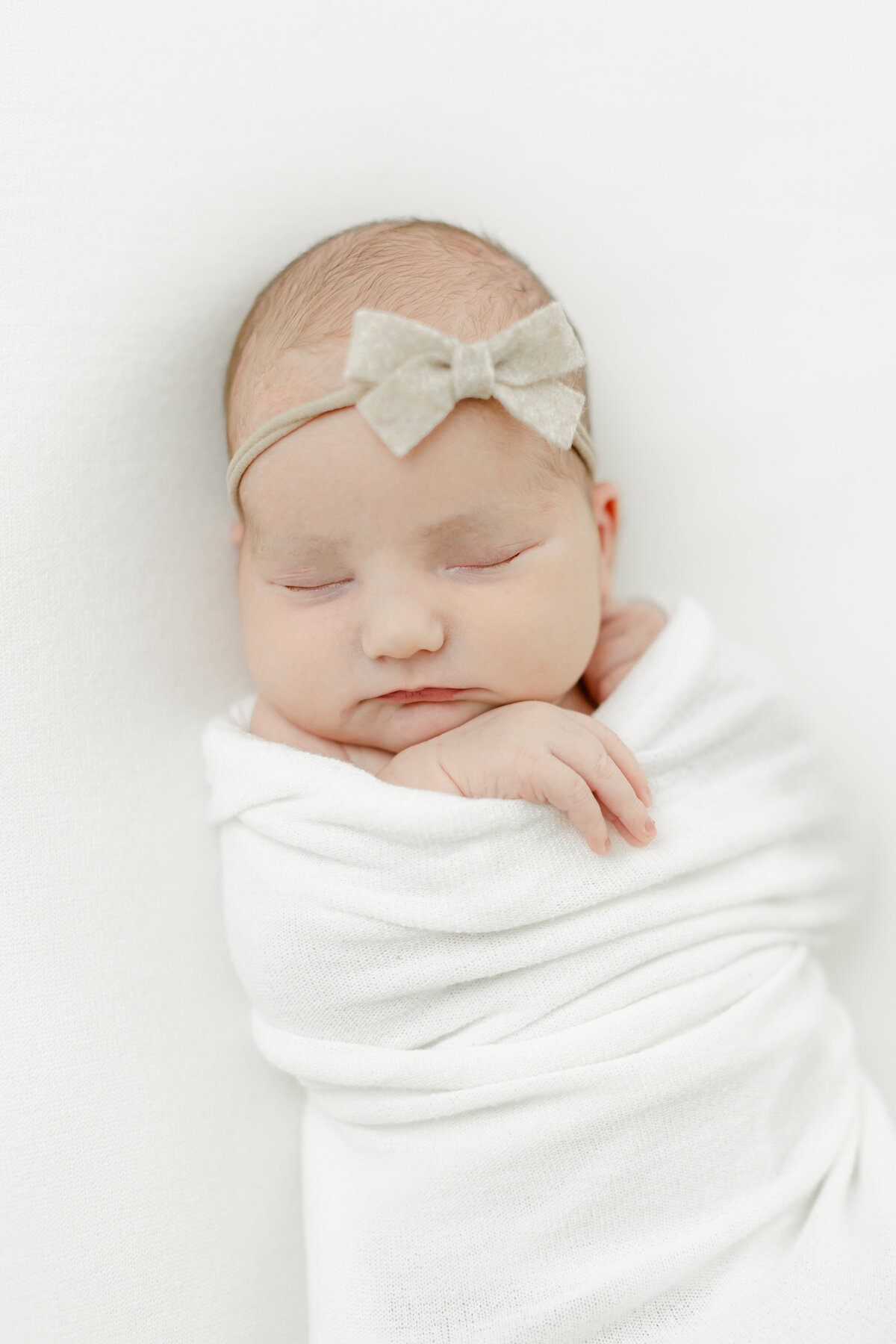 sweet baby girl swaddled and sleeping peacefully in a white swaddle blanket wearing a bow as she is photographed by Philadelphia Newborn Photographer Tara Federico