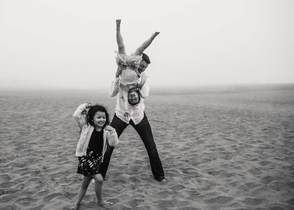 Playful lifestyle photo of dad hanging daughter upside down while other child runs on beach