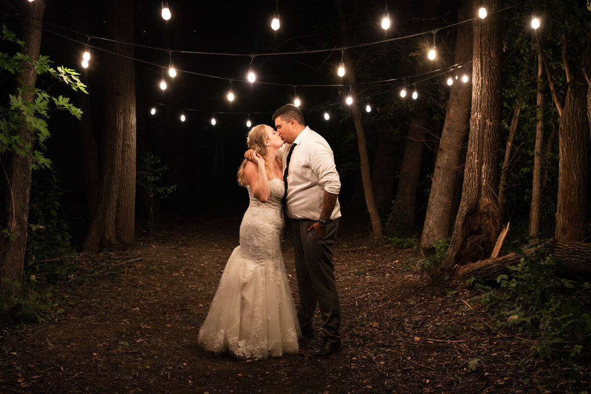 Bride and groom kiss under string lights at night.