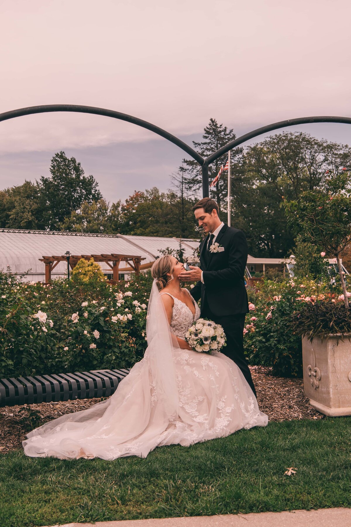 A bride and groom sharing a moment under an archway in a garden, with the bride seated and holding a bouquet, carefully planned by a wedding coordinator in Iowa.