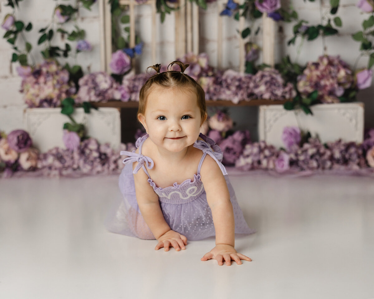 This little cutie is all smiles in a lavender dress, crawling amidst a beautiful purple flower backdrop. Captured by Yucaipa cake smash photographer Kaitlyn Dawn Photography.