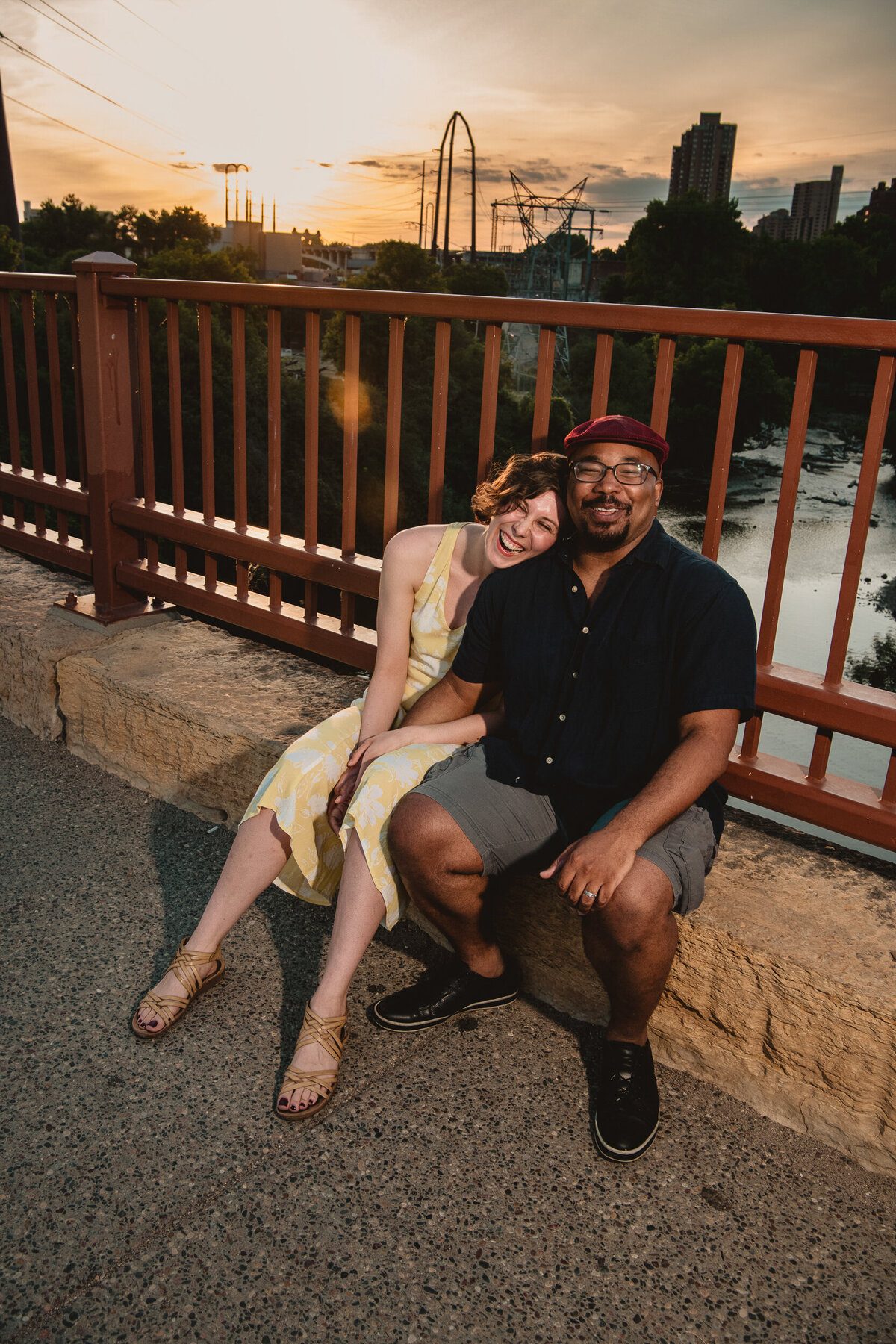 Black man and woman laugh while sitting on the Stone Arch Bridge at sunset.