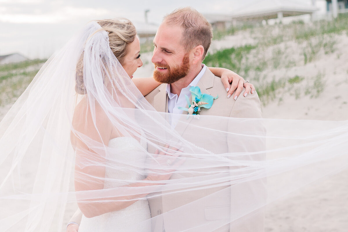 A loving bride and groom on the beach at sunset smiling at each other under a wedding veil enjoying their Raleigh wedding photos
