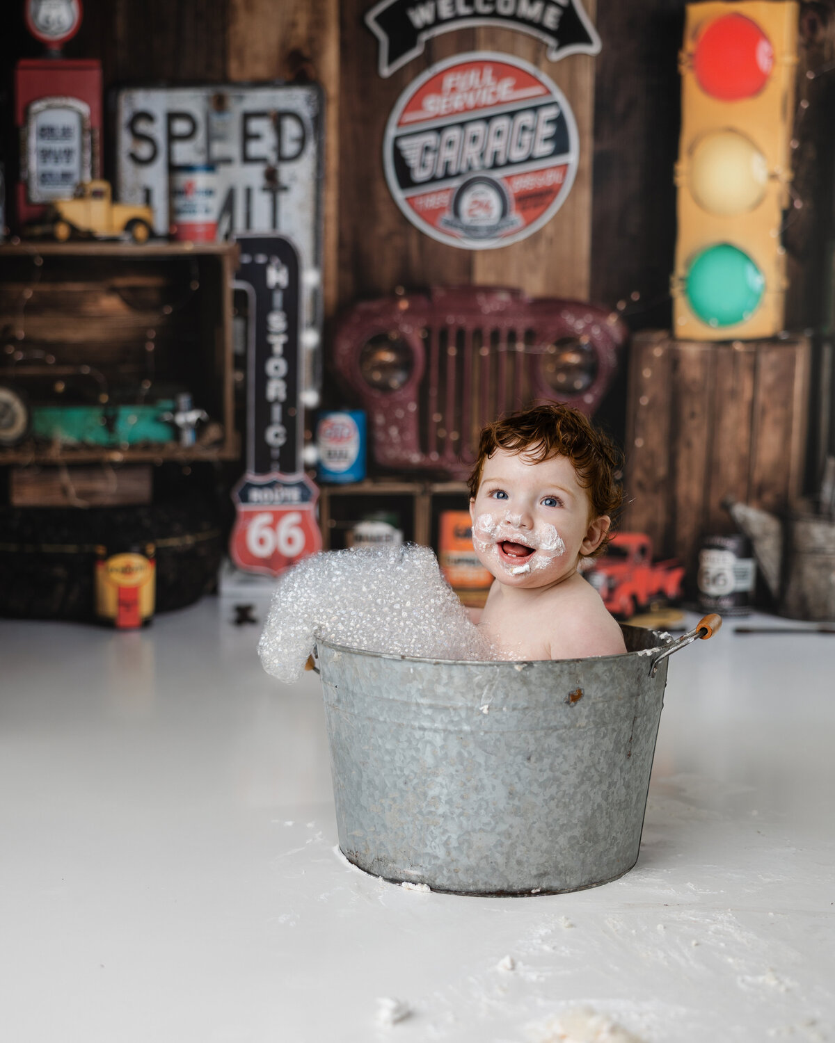 Laughing baby in a bubble bath with frosting on his face, surrounded by retro signs and a rustic truck backdrop. Photograph by Yucaipa cake smash photographer Kaitlyn Dawn Photography.