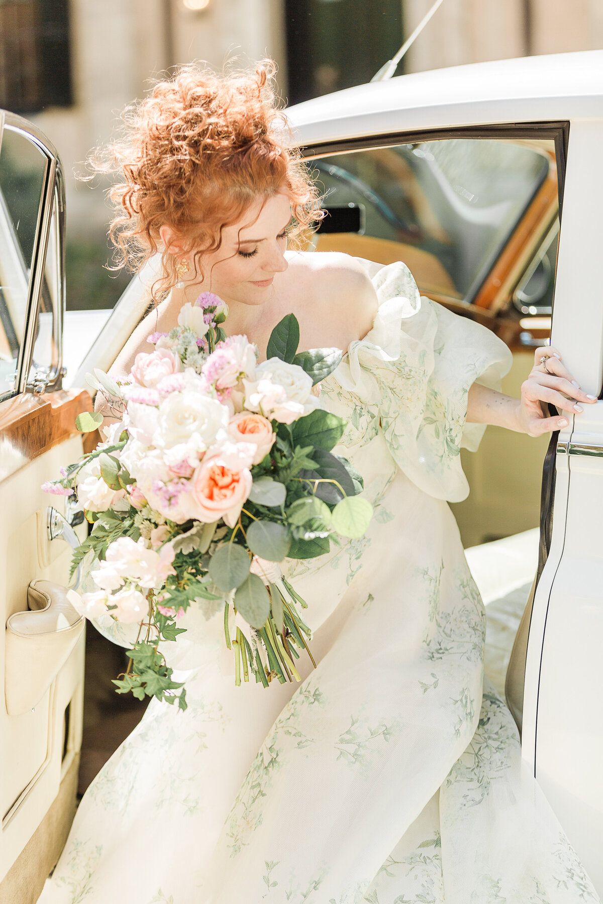 Bride is captured exiting a vintage Rolls Royce holding a large bouquet of lowers. Her red curly hair is upswept. Captured by best Massachusetts wedding photographer Lia Rose Weddings