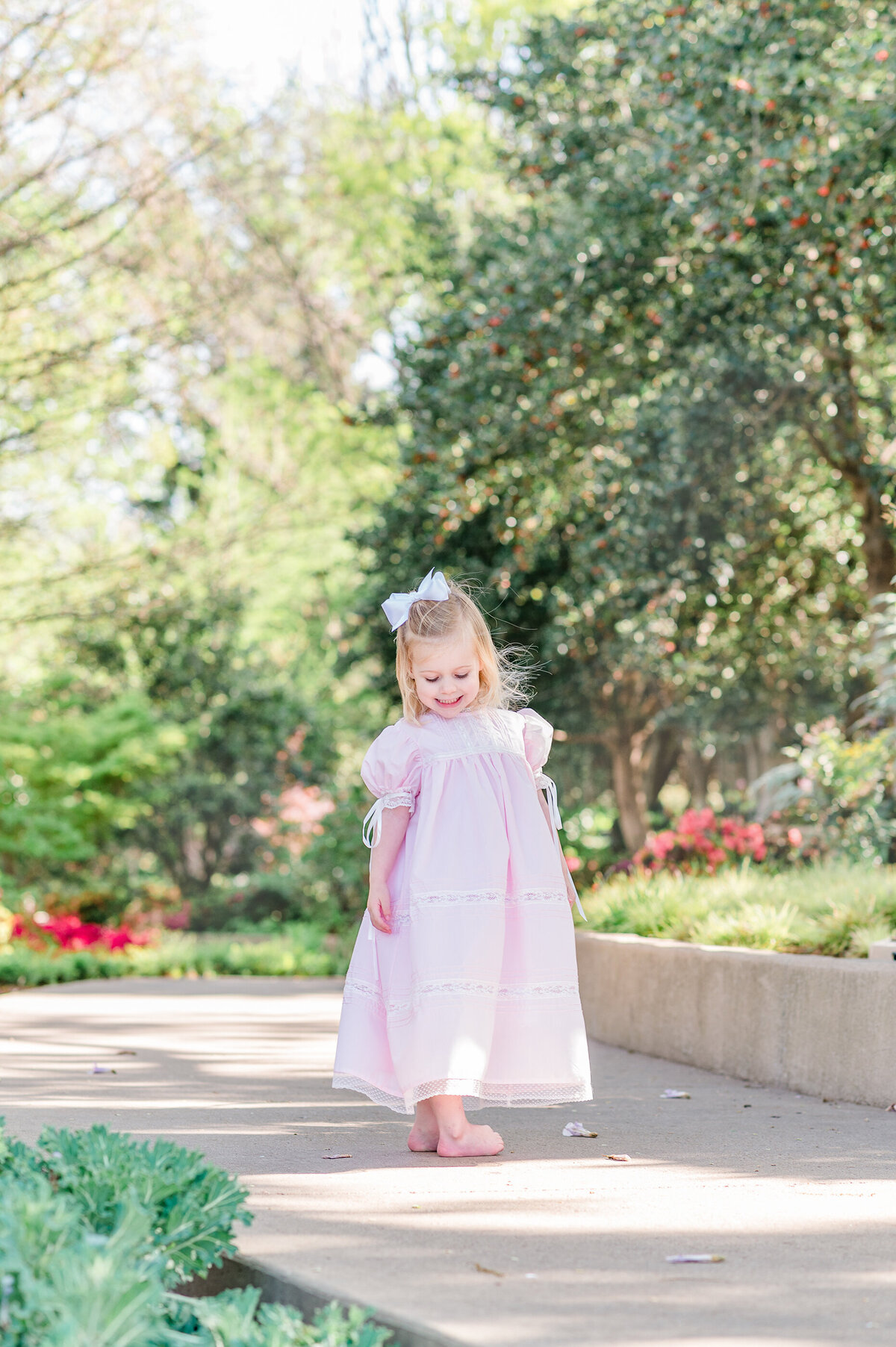 Young girl wearing a pink heirloom dress and large white bow walking down a manicured bath at a botanical garden.