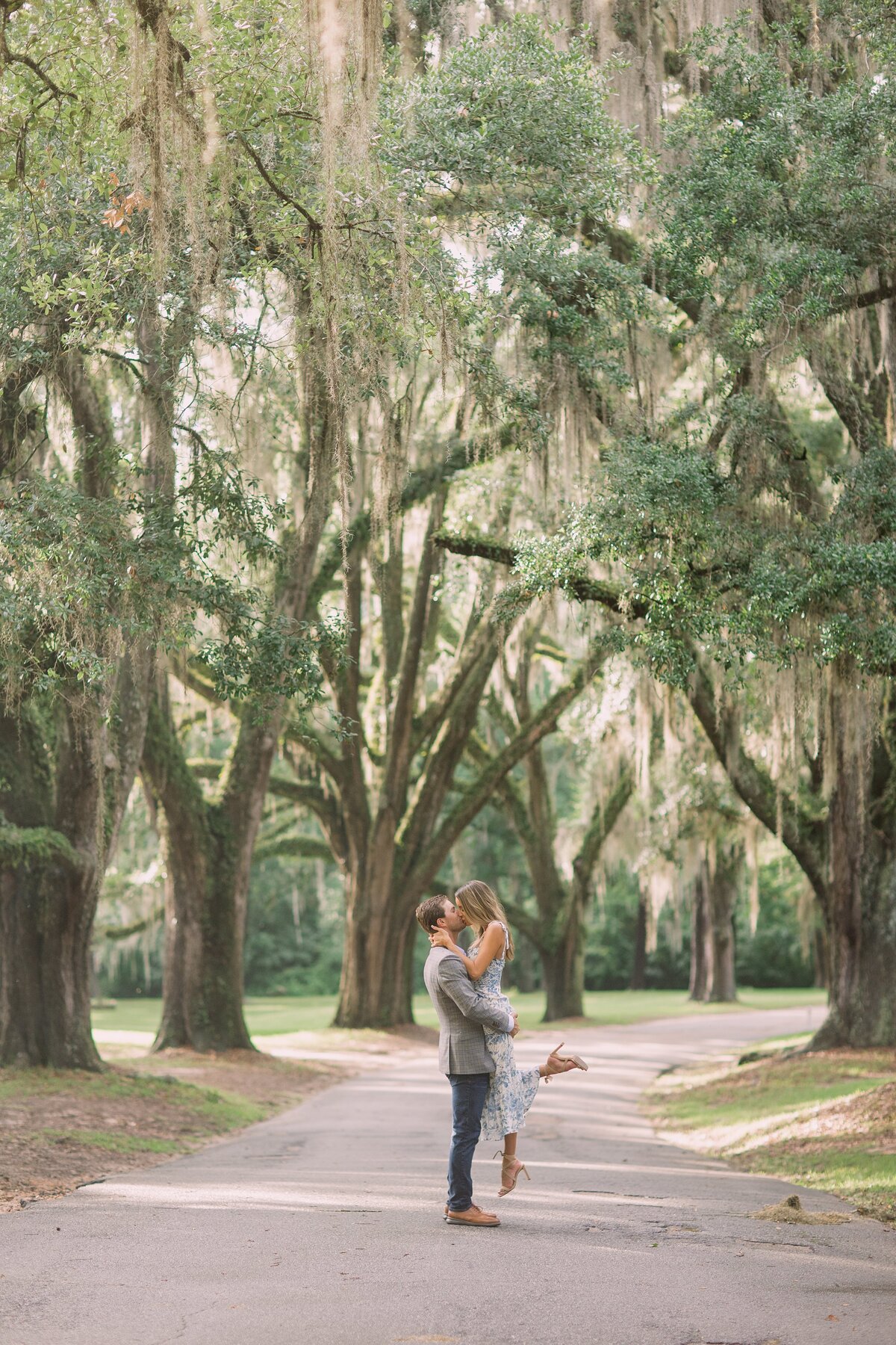 man lifts his fiance up for a kiss on a tree lined path