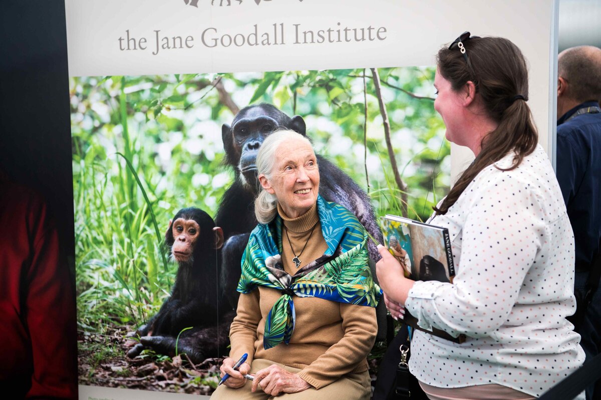 Jane Goodall smiling at a women holding a book with poster of chimpanzees behind her