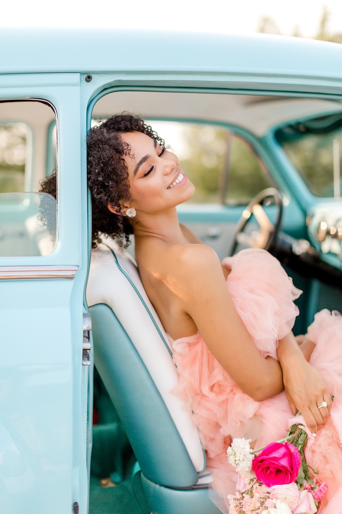 Woman in pink dress sits in the passenger seat of classic car while holding flowers.