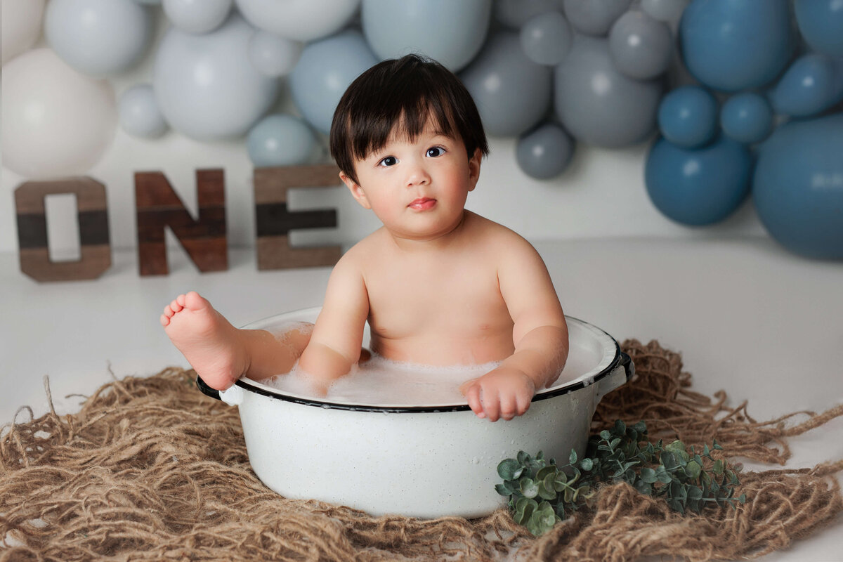 one year old boy sitting in a bubble bath in front of blue balloons and brown one letters at a cake smash session